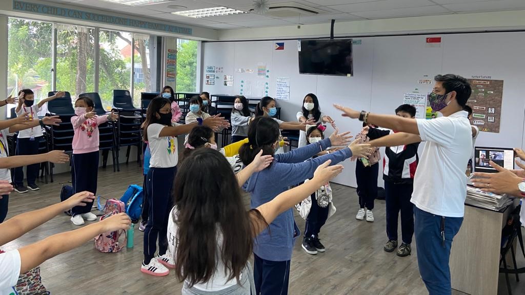 International students in Singapore and their teacher, standing with arms extended forward as part of an activity or exercise.