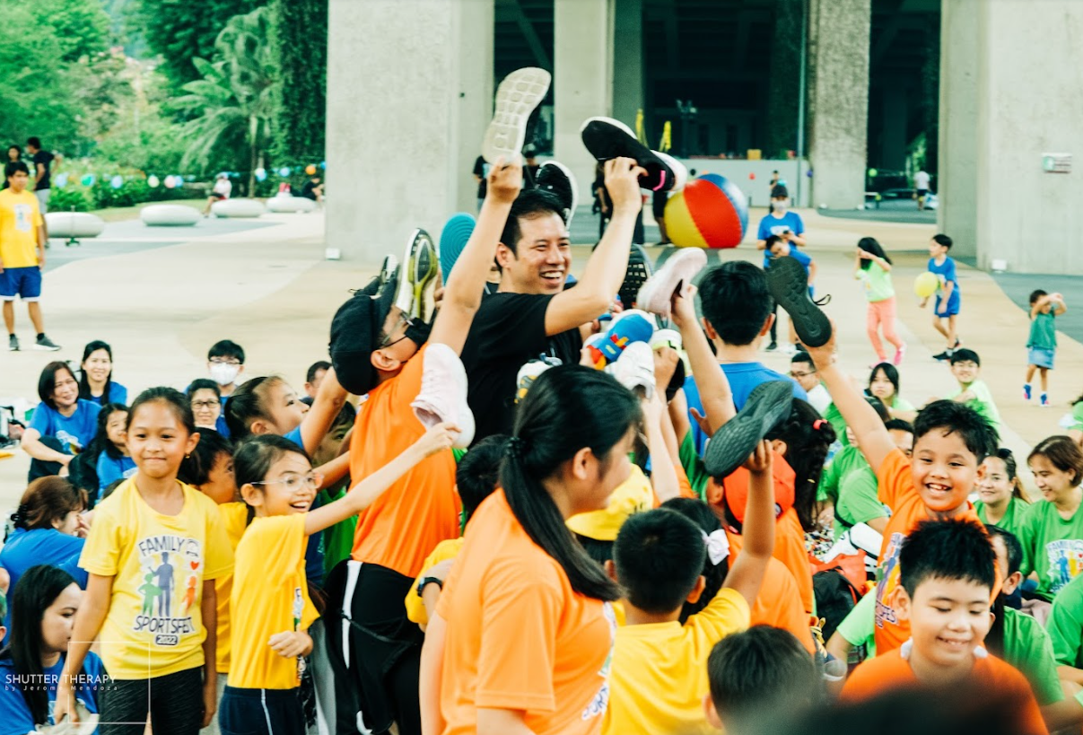 A group of students at a family sportsfest event with their teacher, holding up their shoes.